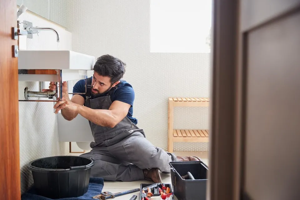 Plumber fixing pipes under sink in bathroom with tools nearby