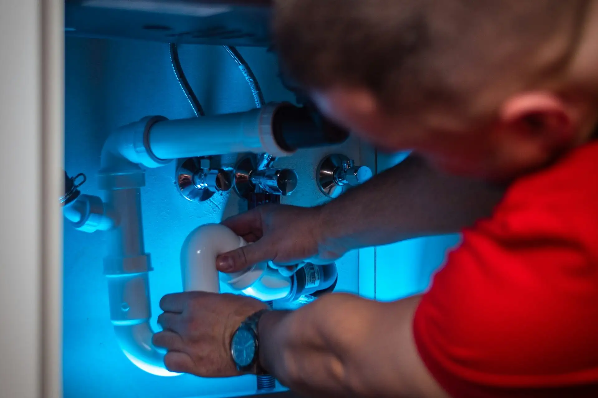 Plumber working on pipes under blue-lit sink with pipe wrench