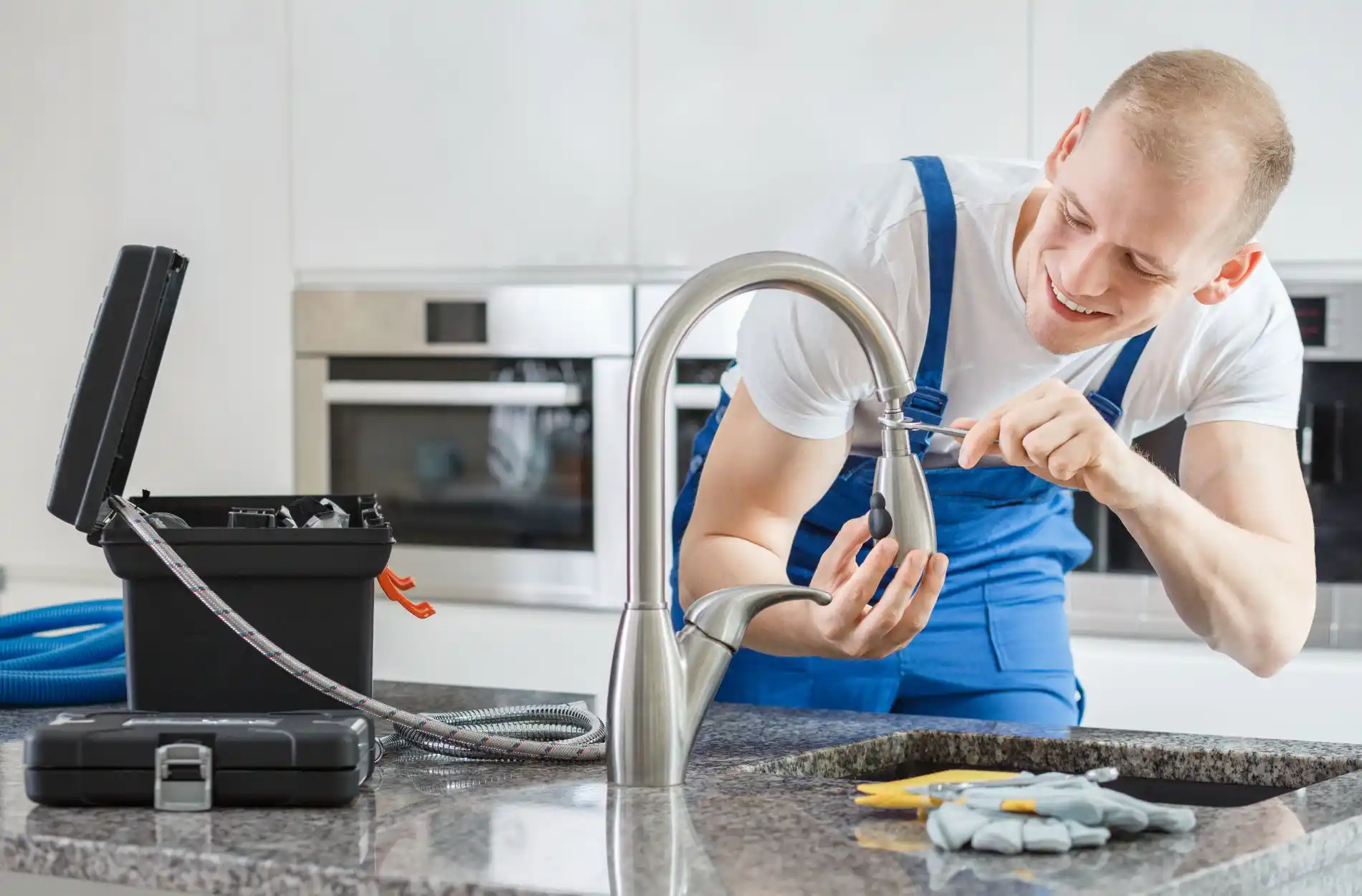 Plumber in blue overalls fixing kitchen faucet with tools nearby