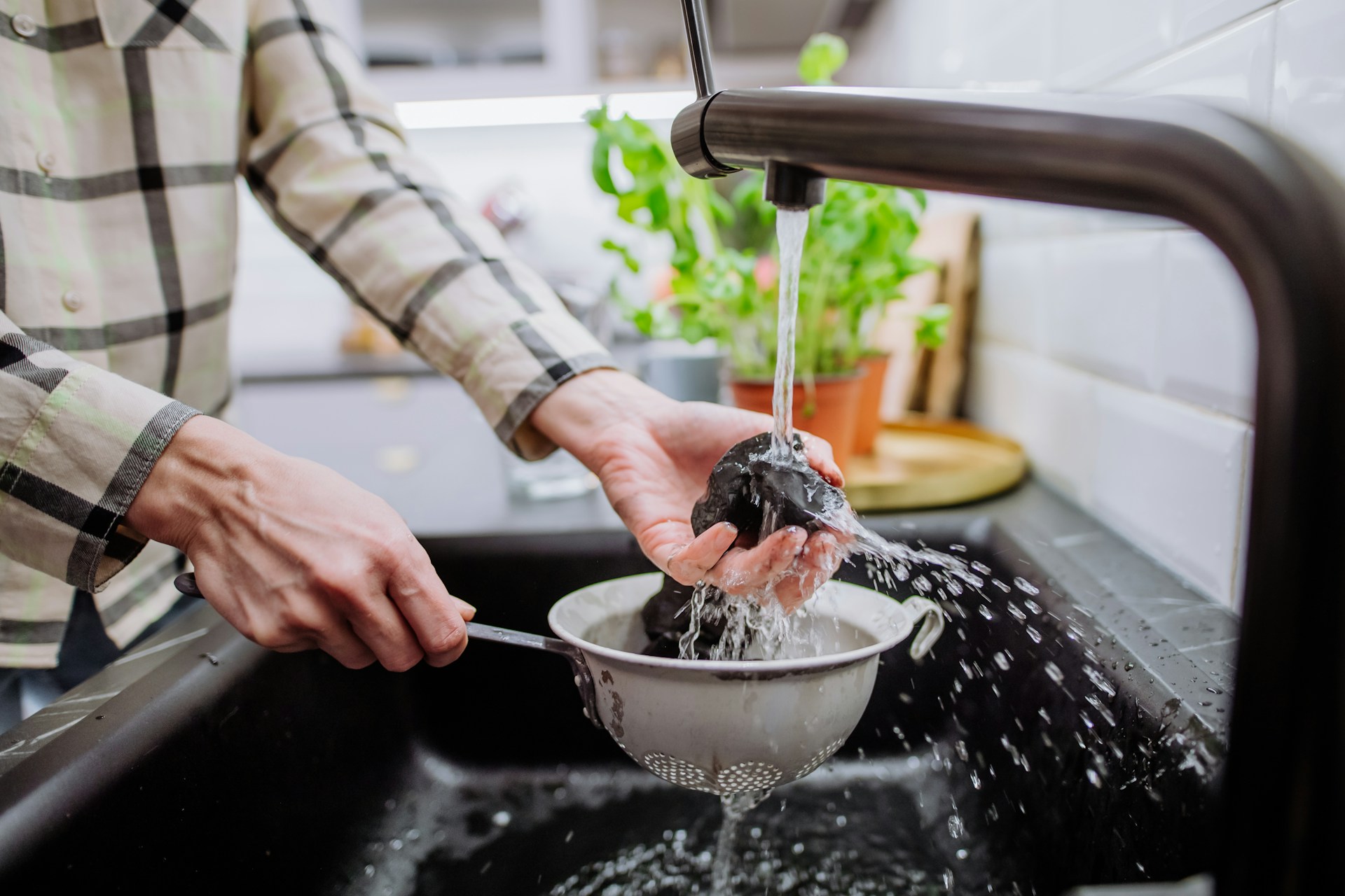 Hands washing vegetables in a colander under running kitchen sink water