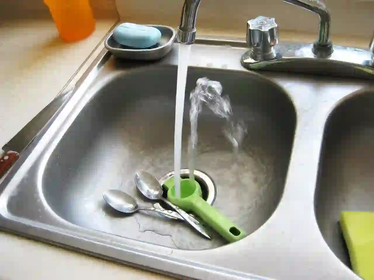 Running water in kitchen sink with spoons and green kitchen tool