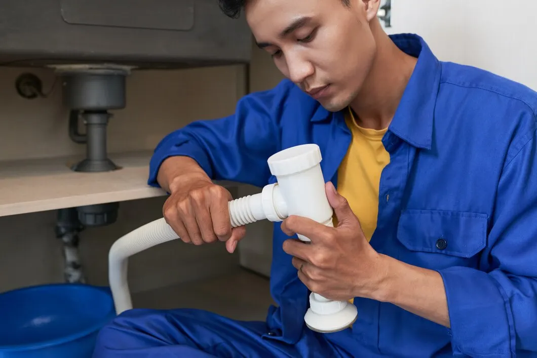 Plumber in blue uniform fixing sink pipe under kitchen counter