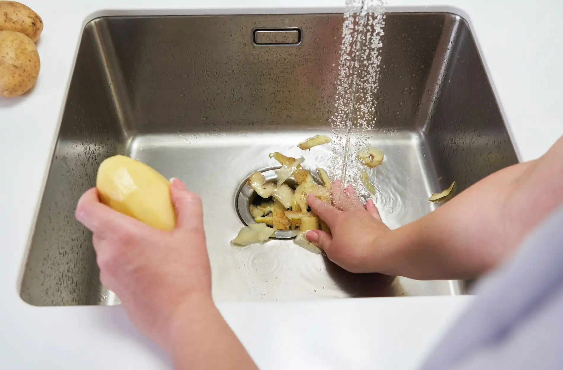 Hands peeling and washing potatoes over a stainless steel kitchen sink