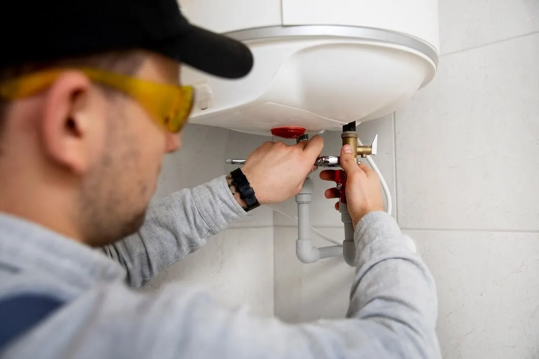 Plumber in safety glasses fixing pipes under a white water heater