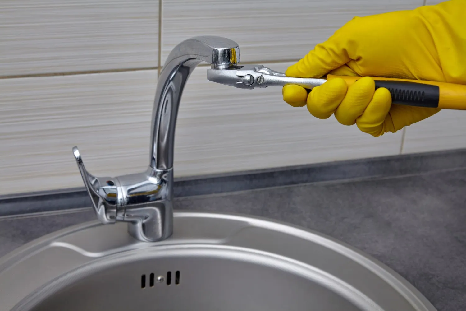 Yellow gloved hand using wrench to repair kitchen sink faucet