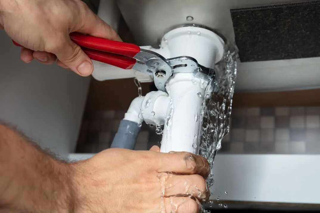 Plumber using wrench to repair leaking white PVC pipe under sink