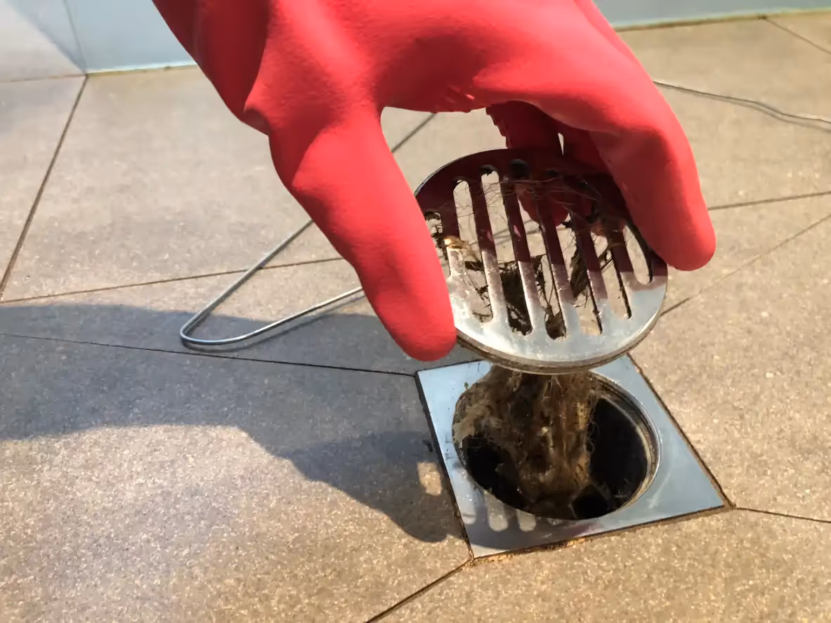 Red gloved hand cleaning hair-clogged drain with metal strainer