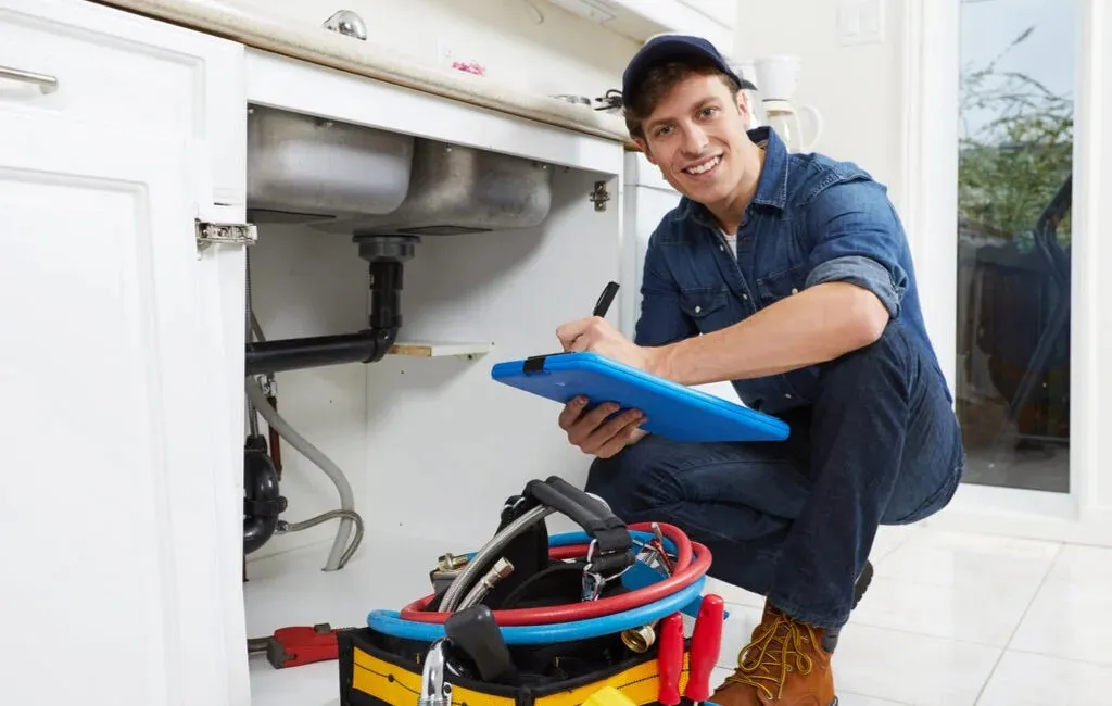Smiling plumber checking pipes under sink with clipboard and tools