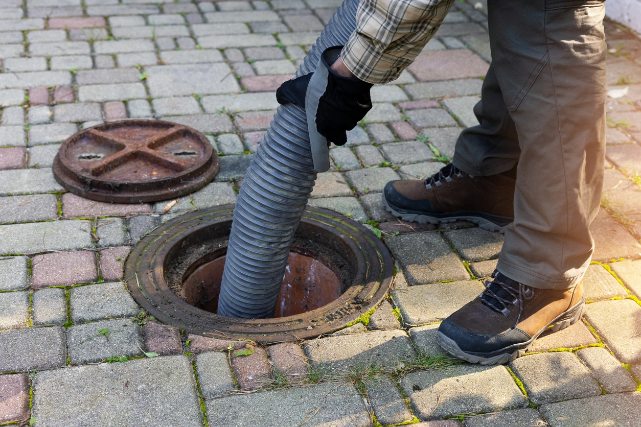 Person in work boots and gloves operates a large hose to vacuum a sewer, surrounded by brick paving. 