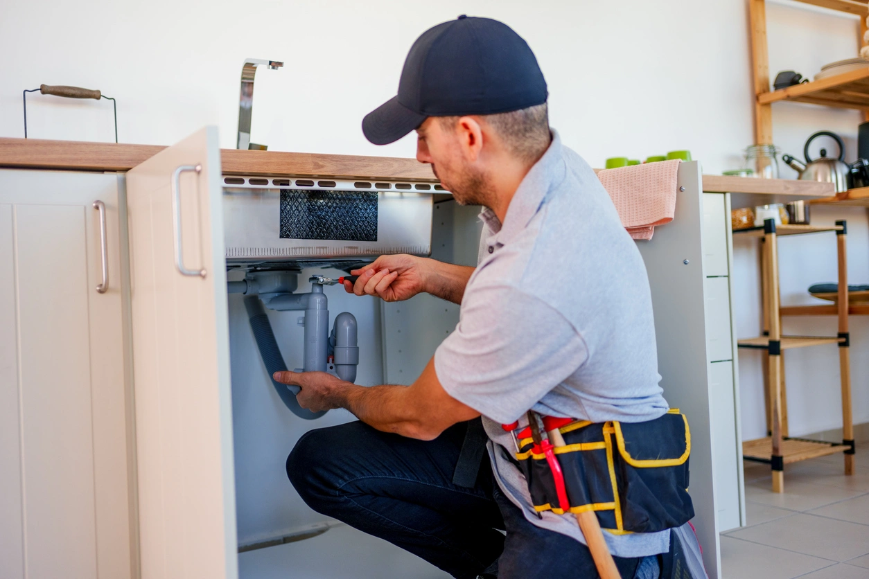Man in a cap fixing kitchen sink pipes under an open cabinet.