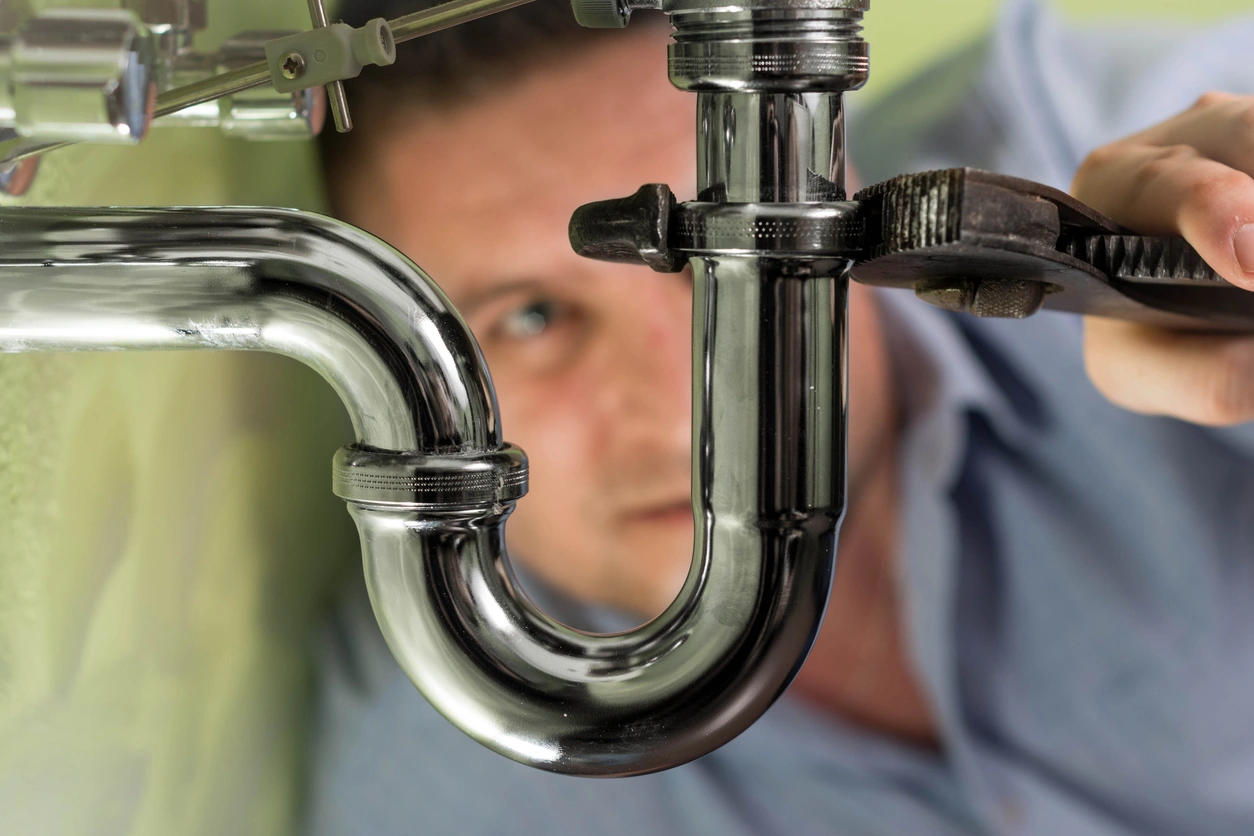 A person uses a wrench to tighten a shiny chrome pipe under a sink, focusing intently. 