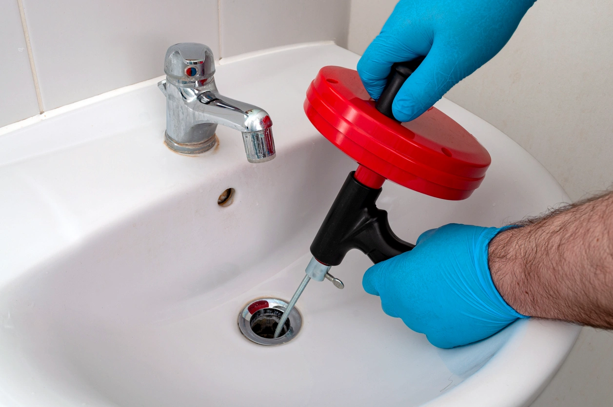 Person wearing blue gloves uses a red drain snake to unclog a white bathroom sink with a chrome faucet. The scene conveys a task-focused mood.