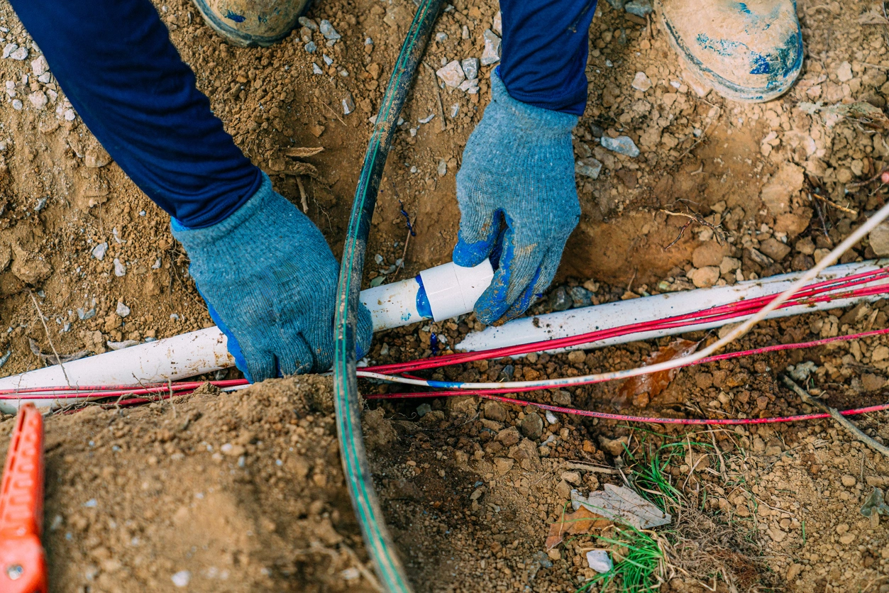 Worker wearing blue gloves and boots repairs a white PVC pipe in a dirt trench, surrounded by red and green cables, conveying precision and focus.