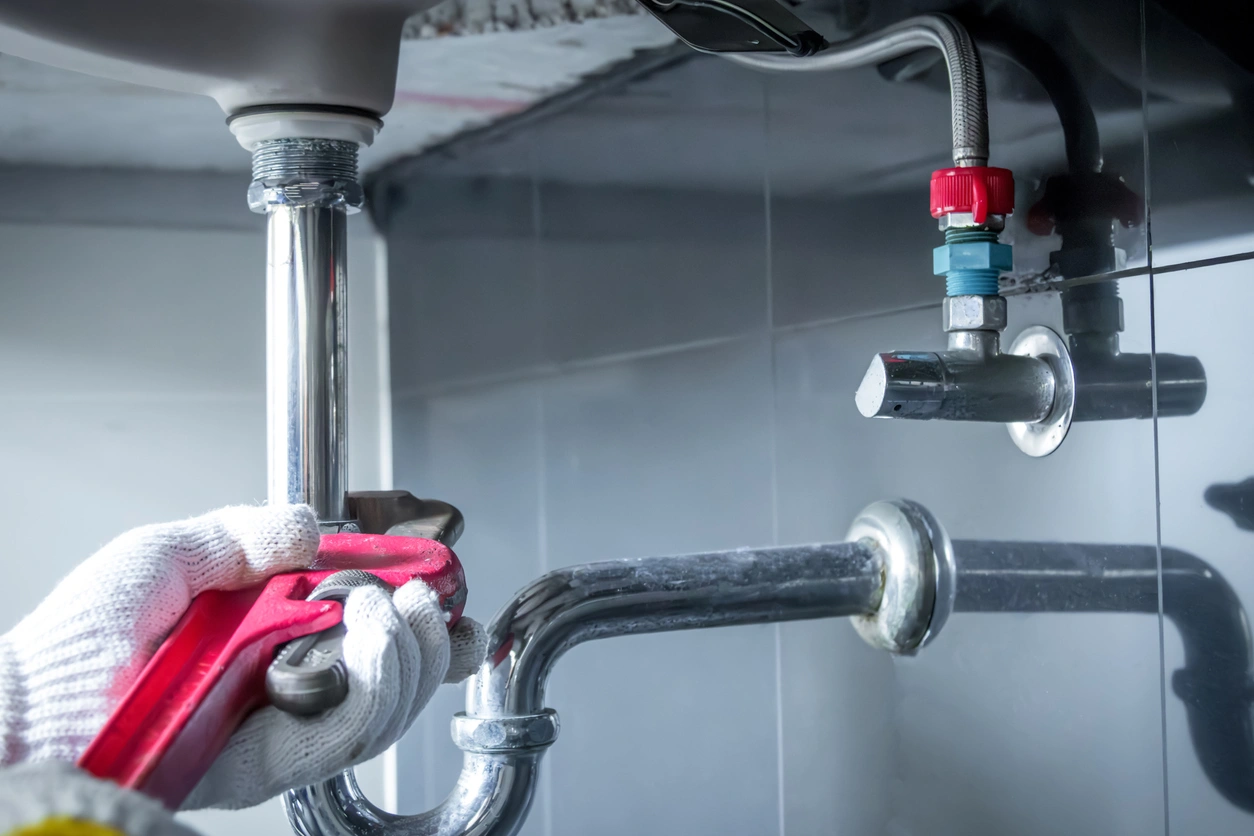 A plumber in white gloves uses a red wrench to fix silver pipes under a sink,