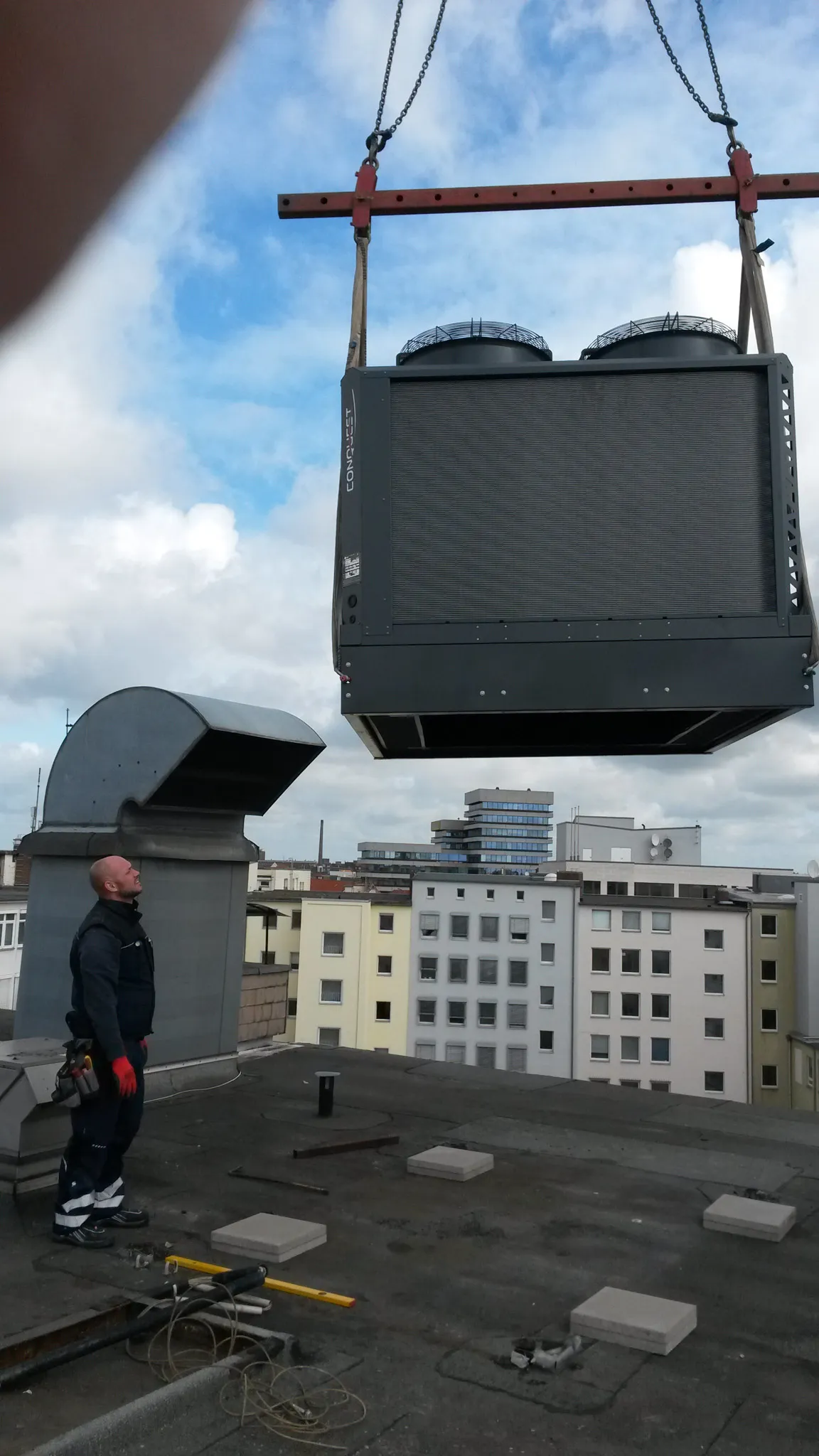 Ein Arbeiter auf einem Flachdach beobachtet, wie ein großes schwarzes Lüftungsgerät mit einer Kranvorrichtung angehoben wird.