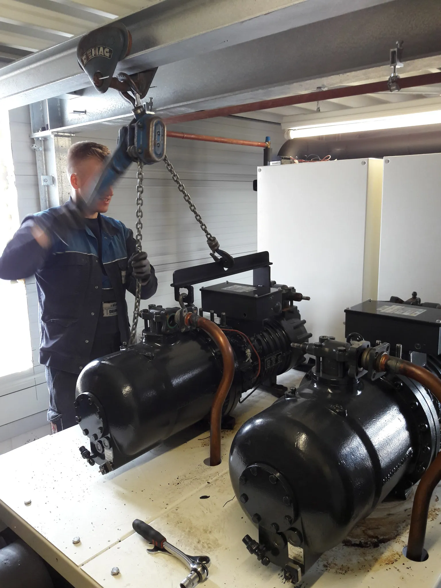 Techniker hebt mit Kettenzug einen schwarzen Kompressor in einer Werkstatt.