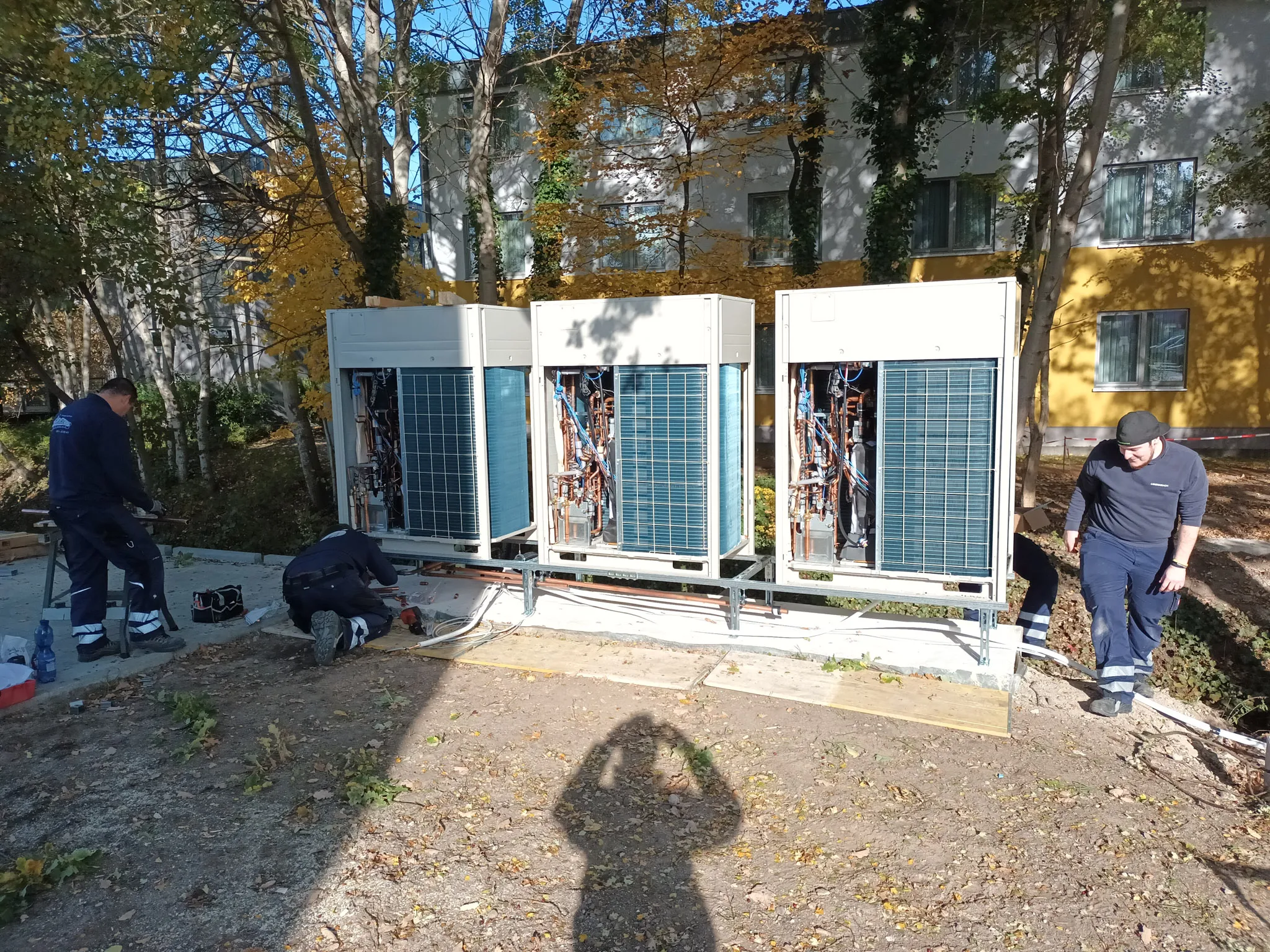 Drei Techniker arbeiten an großen Klimageräten im Freien neben einem Gebäude mit gelb-weißer Fassade.