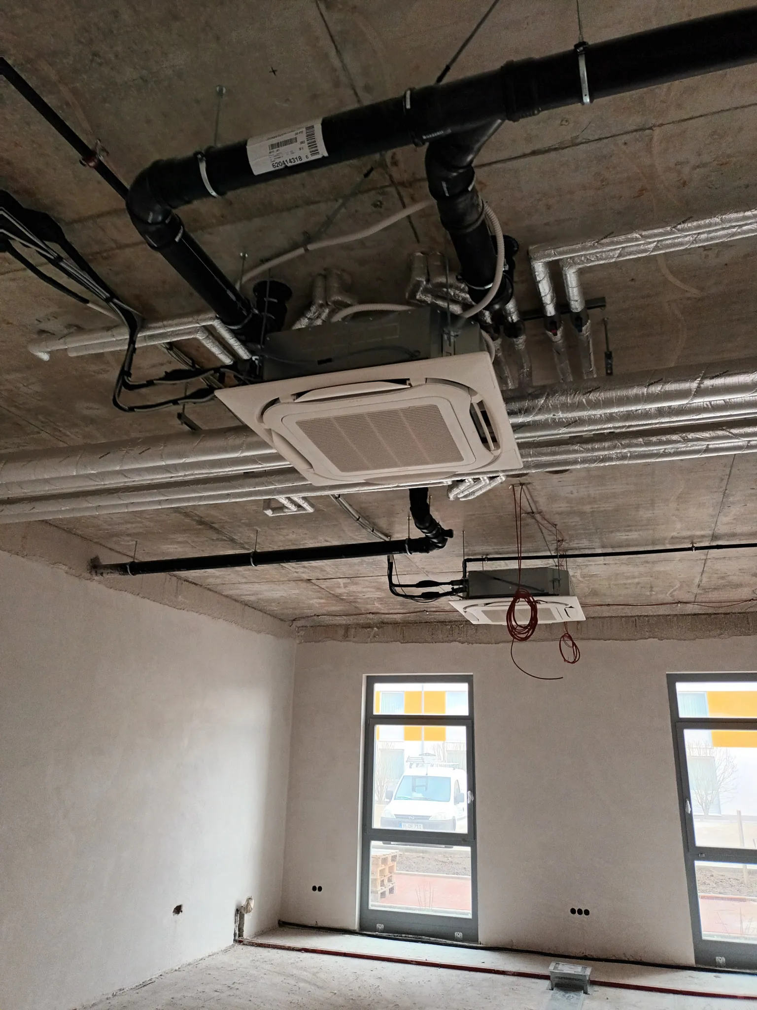 Innenraum eines unfertigen Raums mit sichtbaren Rohrleitungen und installierten Klimageräten an der Decke.