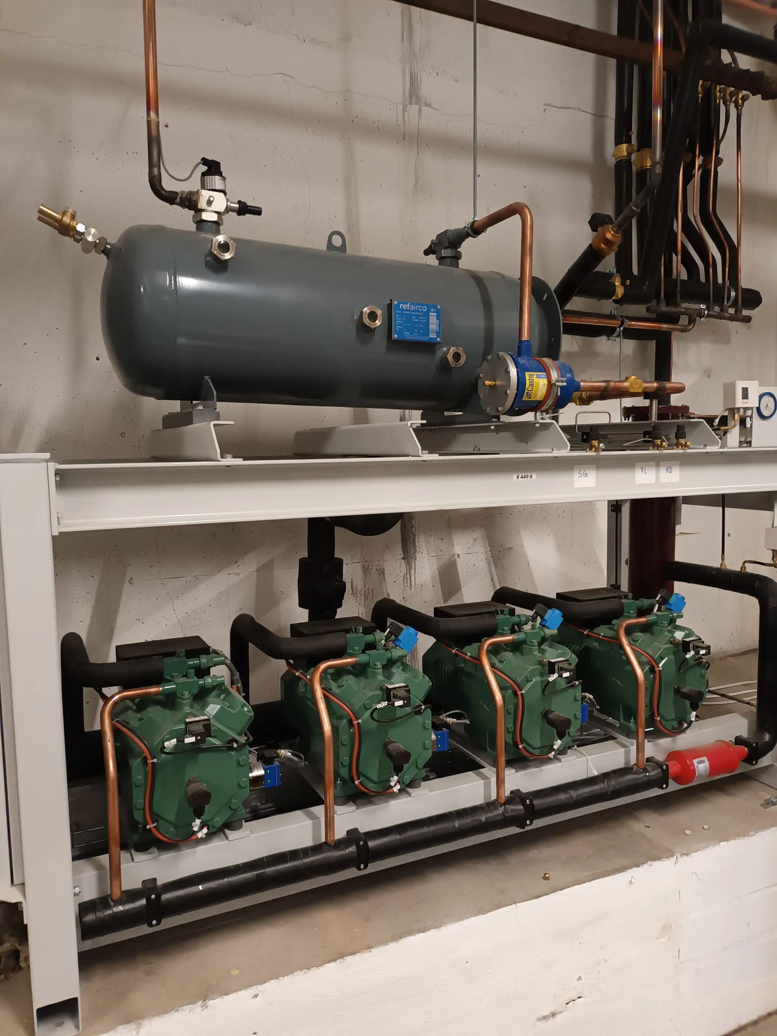 Industrielle Kälteanlage mit vier grünen Kompressoren und einem großen grauen Luftbehälter, verbunden durch Kupferrohre.