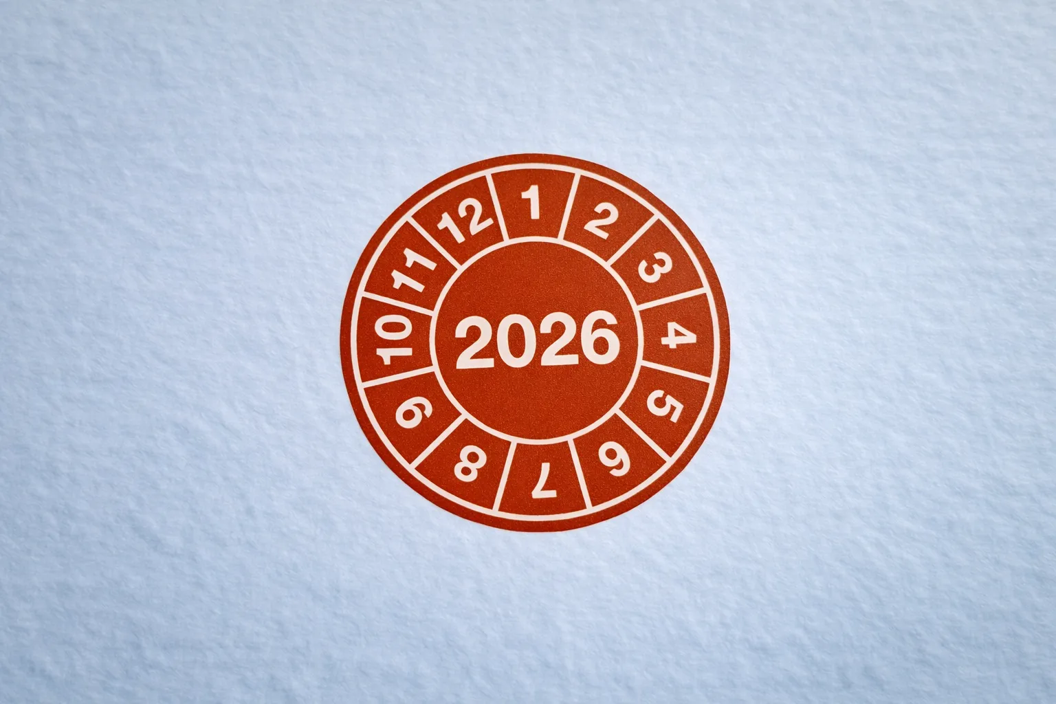 Rote Prüfplakette mit der Jahreszahl 2026 und den Monaten 1 bis 12 in einem Kreis auf weißem Hintergrund.