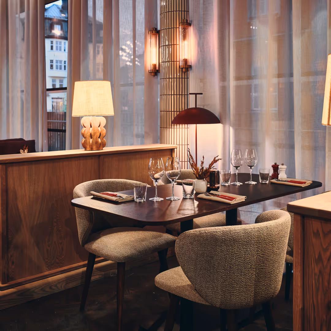 Mysigt restaurangbord med fyra stoppade stolar, dukat med glas, bestick och servetter, omgiven av mjuk belysning och gardiner.