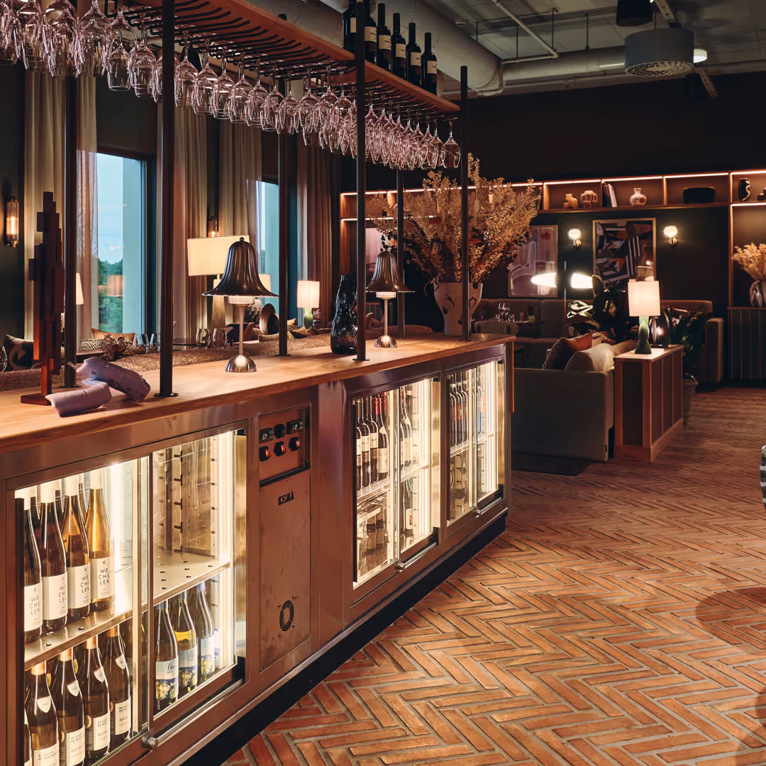 Elegant bar med hängande vinglas, kyld förvaring med vinflaskor och mjukt belysta sittområden med växter och konstverk.