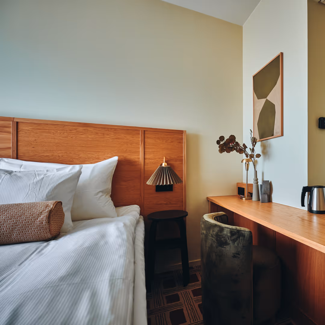 Sovrum med säng med vit sängkläder, träpanelsänggavel, svart sidobord, grön sammetstol och skrivbord med vas och vattenkokare.