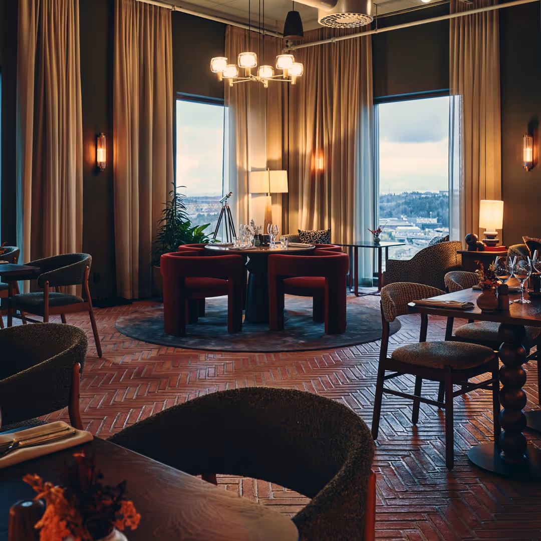 Elegant restauranginteriör med röda stolar runt ett runt bord, stora fönster med utsikt över stad och gardiner, varma ljuskällor och en lyxig atmosfär.