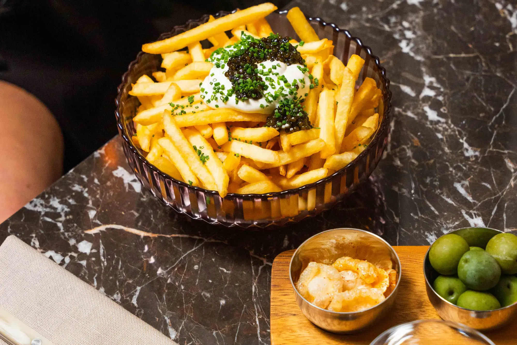 Skål med franska pommes frites toppade med gräddfil, svart kaviar och gräslök på en marmorbordsskiva, bredvid skålar med knapriga snacks och gröna oliver.