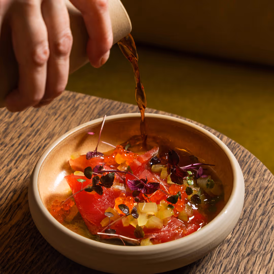 Hand häller en mörk sås över en skål med rå lax, röda röd microgreens och små orange fiskromspärlor.