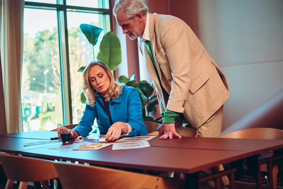 Två affärspersoner som diskuterar och granskar dokument på ett bord i ett kontorsrum med stora fönster i bakgrunden.