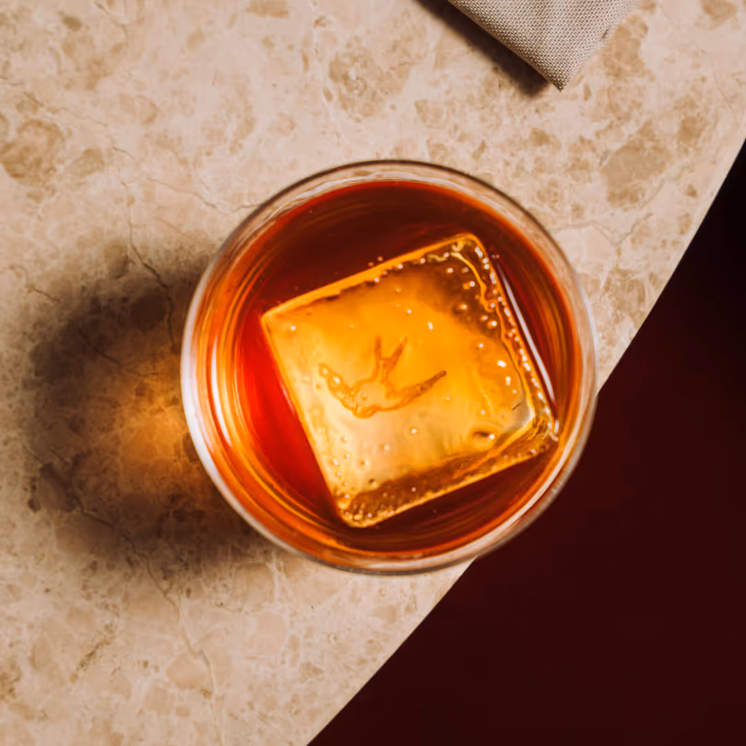 Fågelperspektiv på ett glas med brunt dryck och en stor isbit på en marmorbordsskiva.