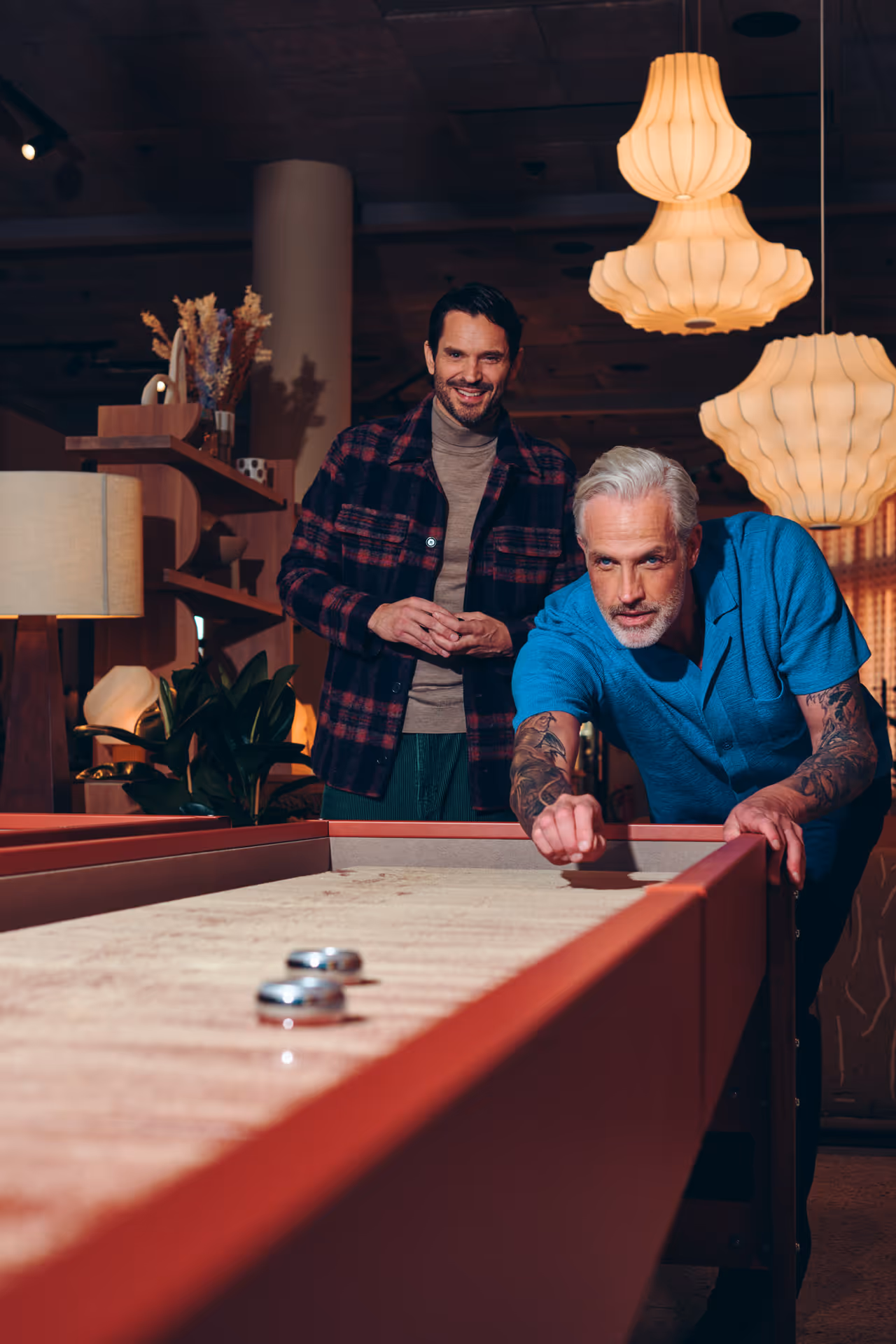 Två män spelar shuffleboard i ett varmt upplyst rum med hängande lampor och trädekor.