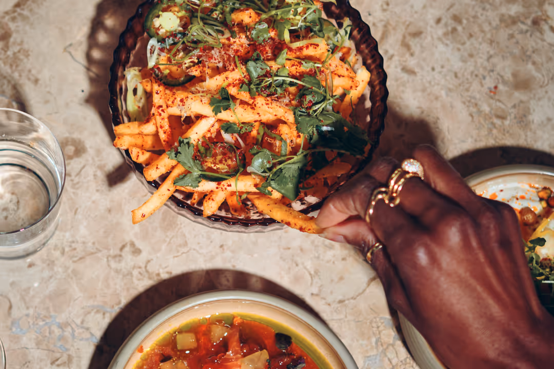 Hand med flera ringar som tar en kryddig pommes frites från en skål med färska örter och kryddor, med ett glas vatten och en tallrik soppa på marmorbord.