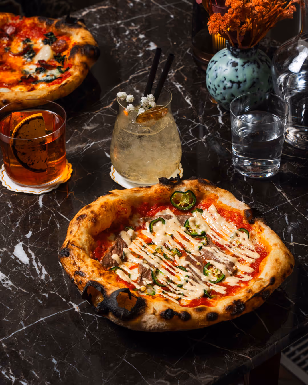 Två pizzor på ett mörkt marmorbord med två cocktails, ett glas vatten och en vas med orange blommor.