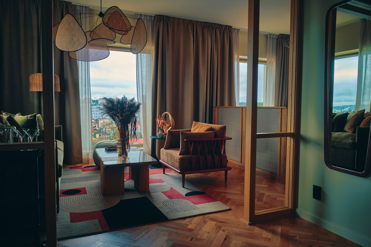 Ett hotellsrum med ett soffbord, en fåtölj, stora fönster med gardiner och en färgglad matta på trägolvet.