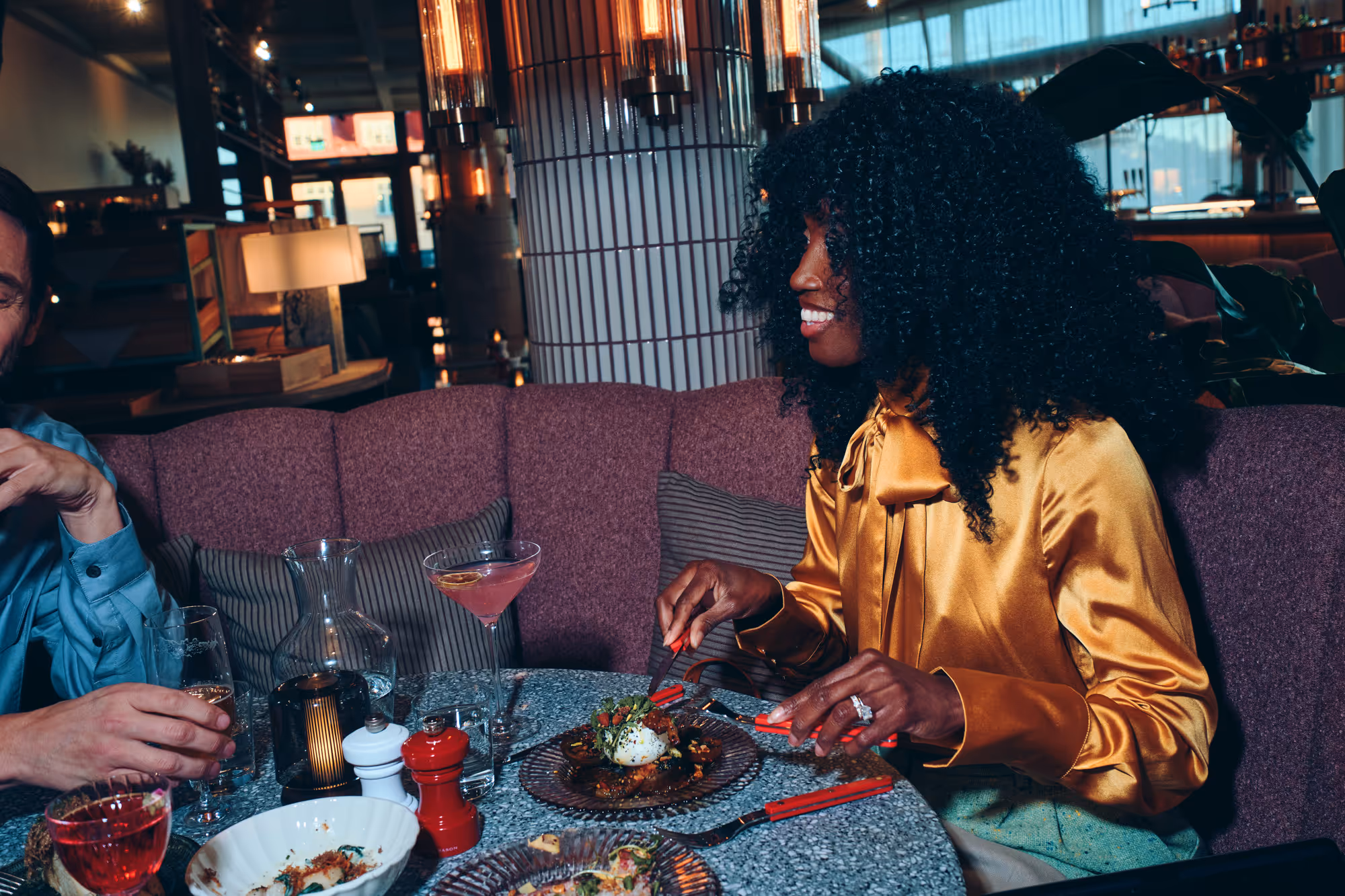 Kvinna i gul blus äter middag och ler vid bord i en mysig restaurang med cocktails och mat på bordet.