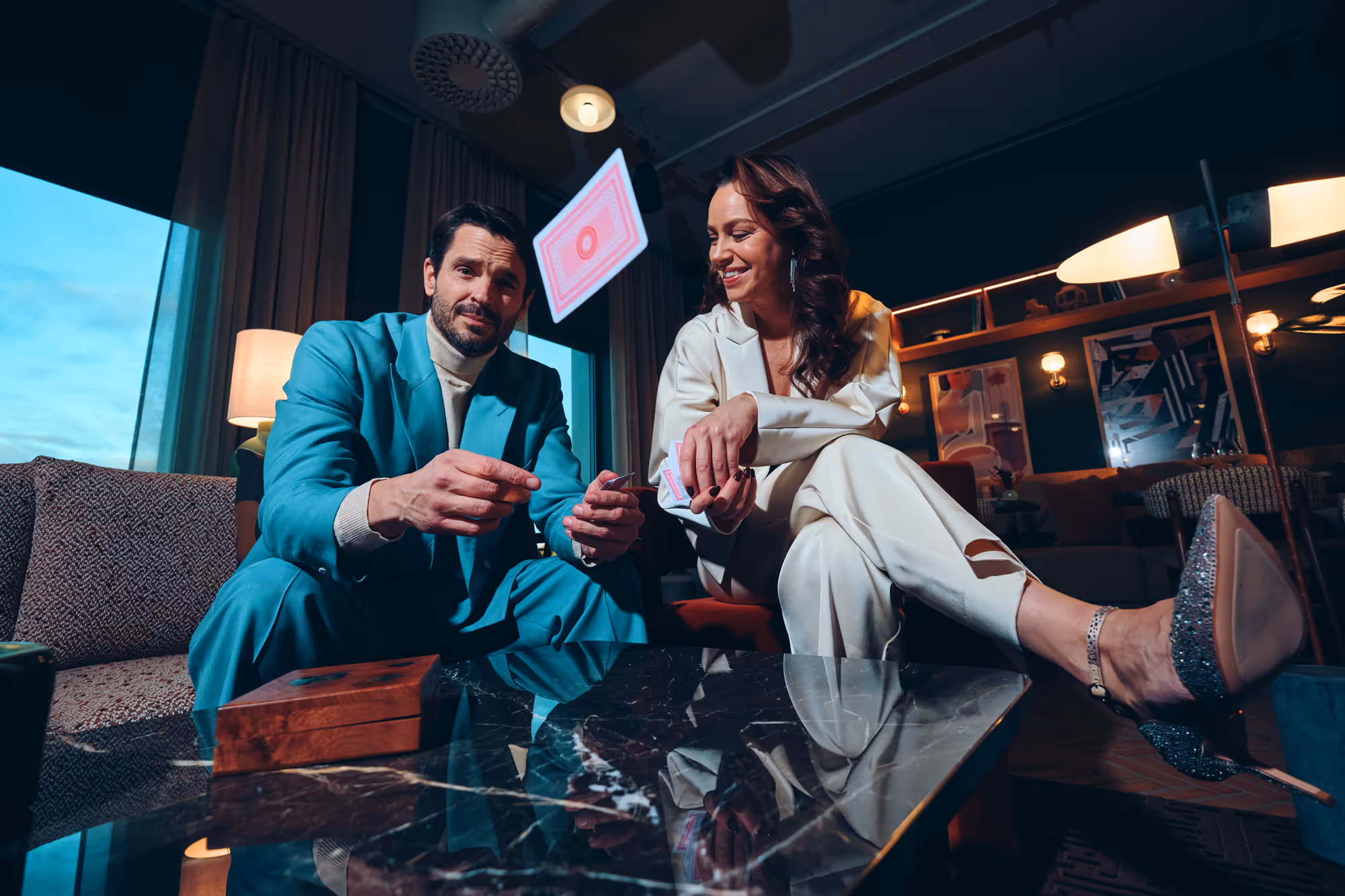 Man i turkos kostym och kvinna i vit kostym spelar kort vid ett marmorbord i ett stilfullt vardagsrum.