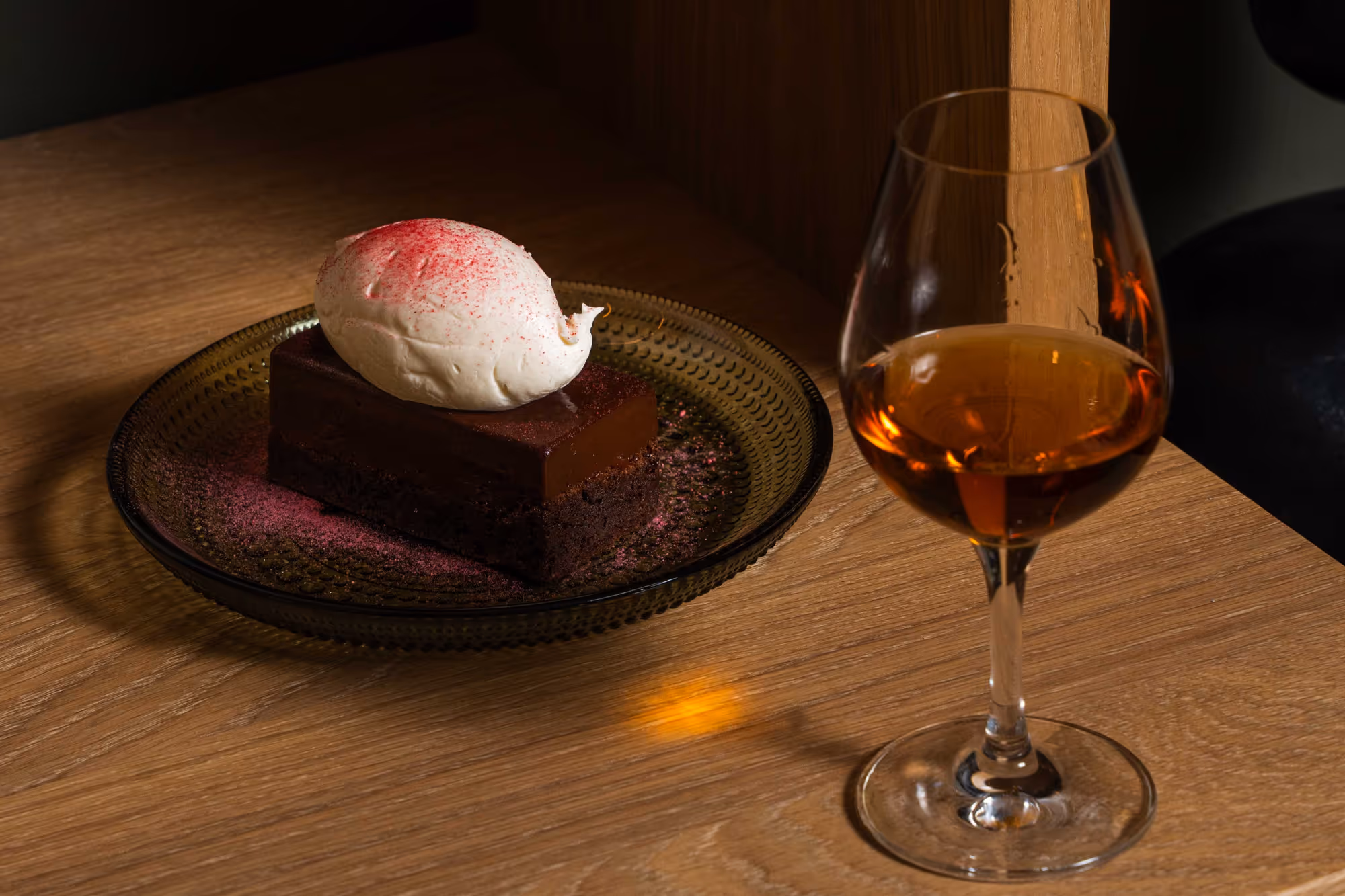 Smörkaka med en klick vispad grädde och rosa pulver på en mörk, mönstrad tallrik samt ett glas ljusbrun likör på ett träbord.