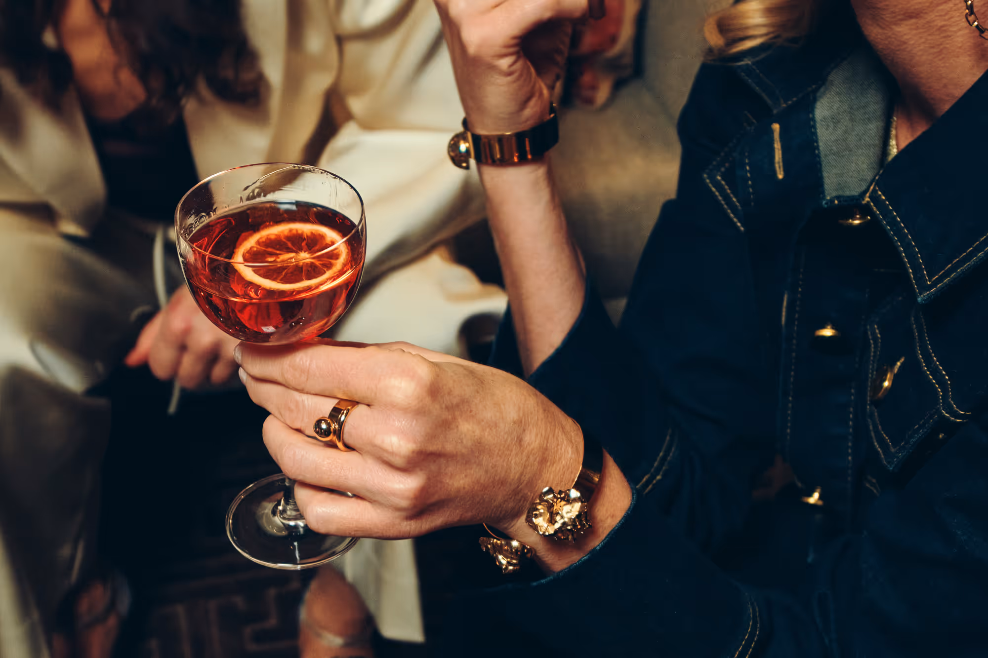 En person håller ett cocktailglas med röd drink och en citronskiva, klädd i mörk jacka med guldarmband och ringar.