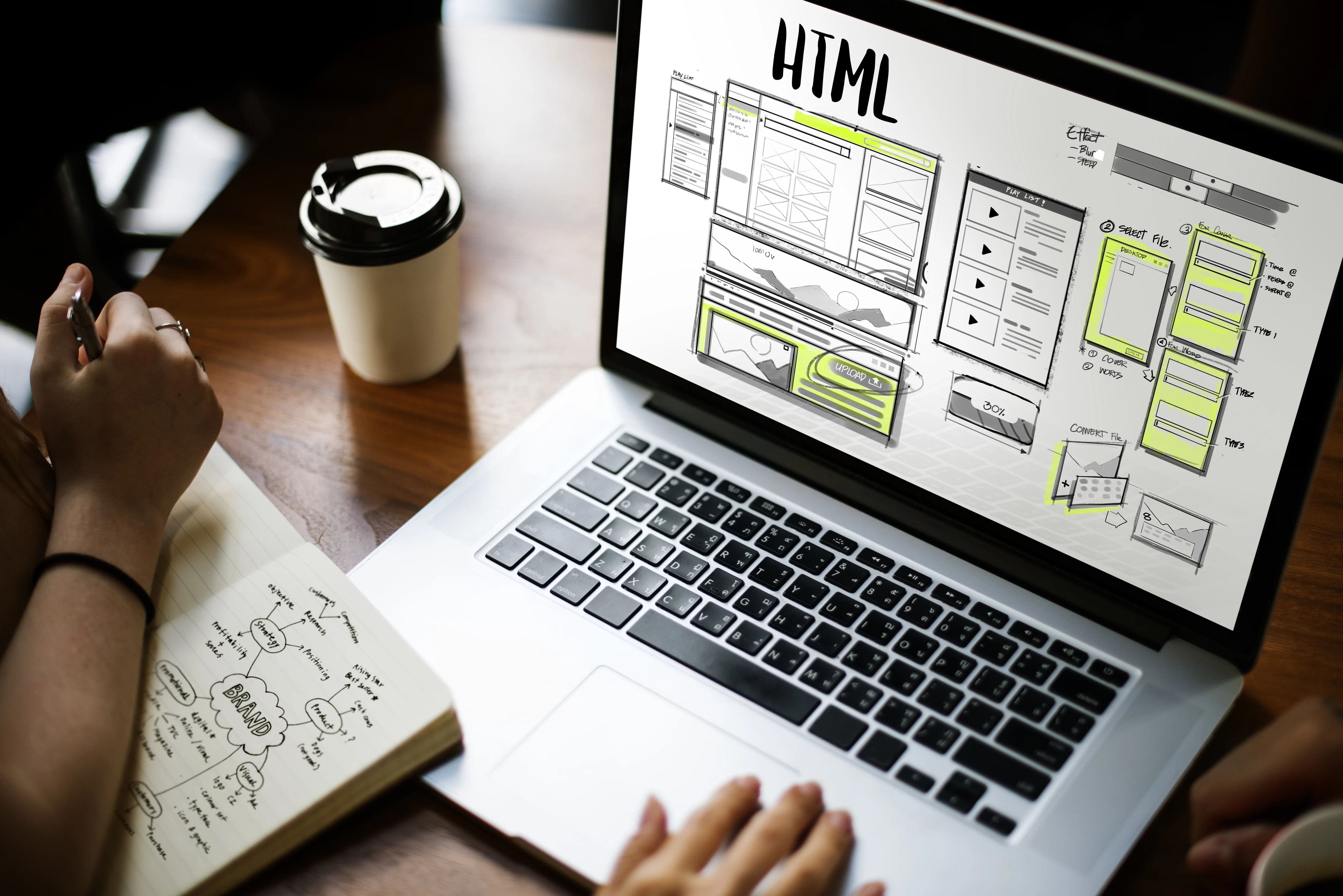 Designer planning website redesign with HTML wireframes on laptop screen and branding notes in notebook.