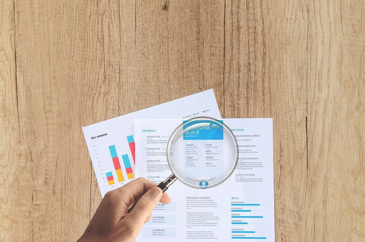 Hand holding magnifying glass over resume and charts related to UX Audit Services on wooden desk.