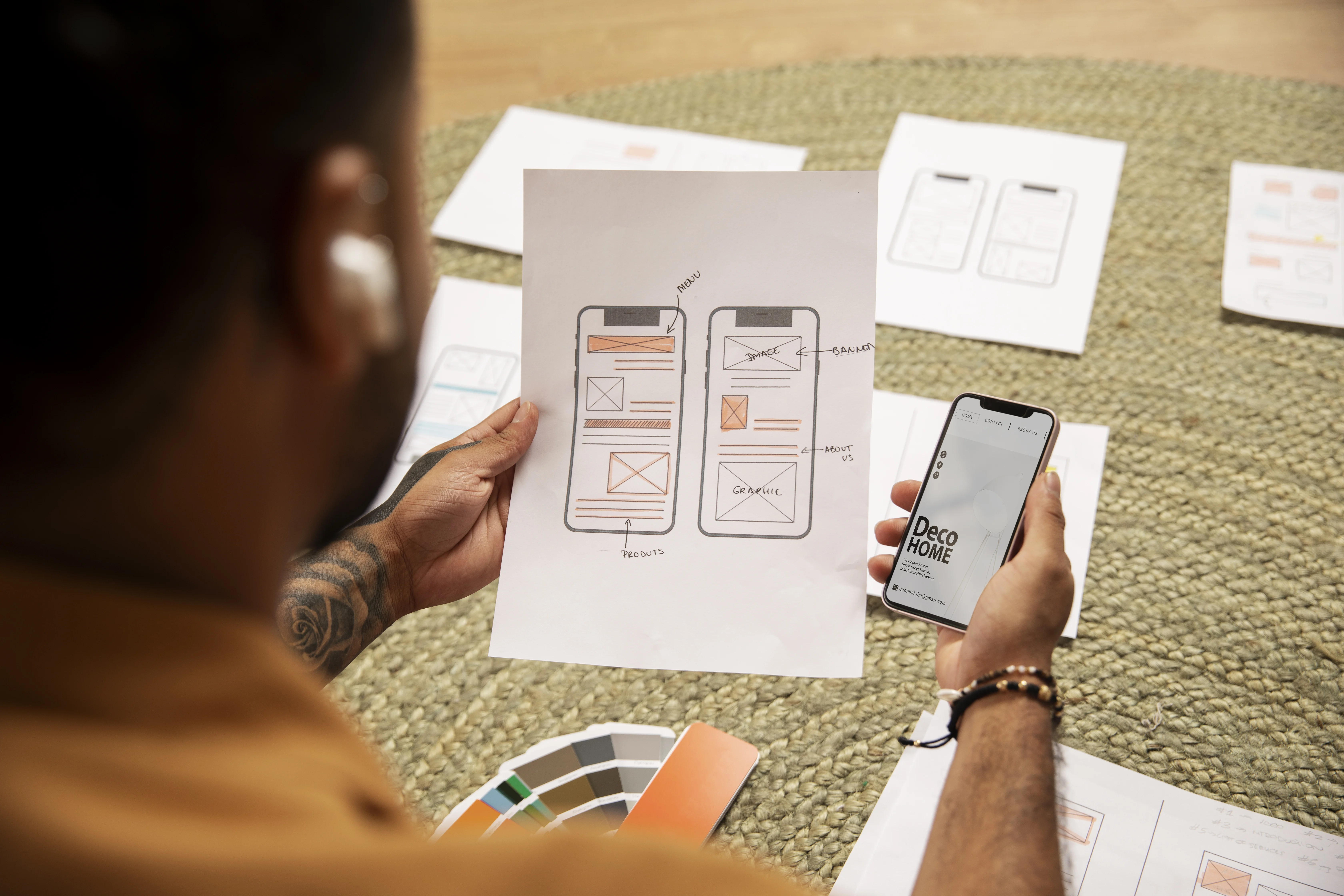 Responsive Design for Mobile Apps: Designer with tattoo holding wireframe sketches and smartphone on rug with color swatches.