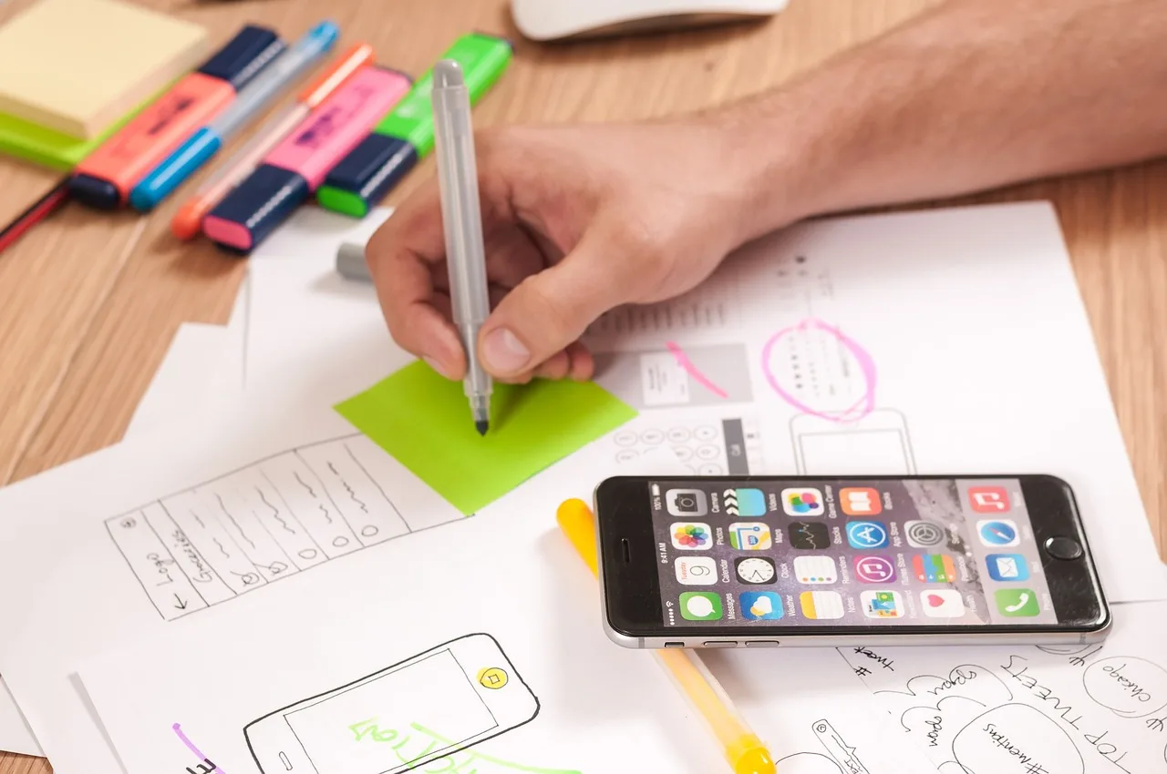 Responsive Design for Mobile Apps: Designer sketching wireframes with pen on paper, smartphone, and colorful markers on desk.