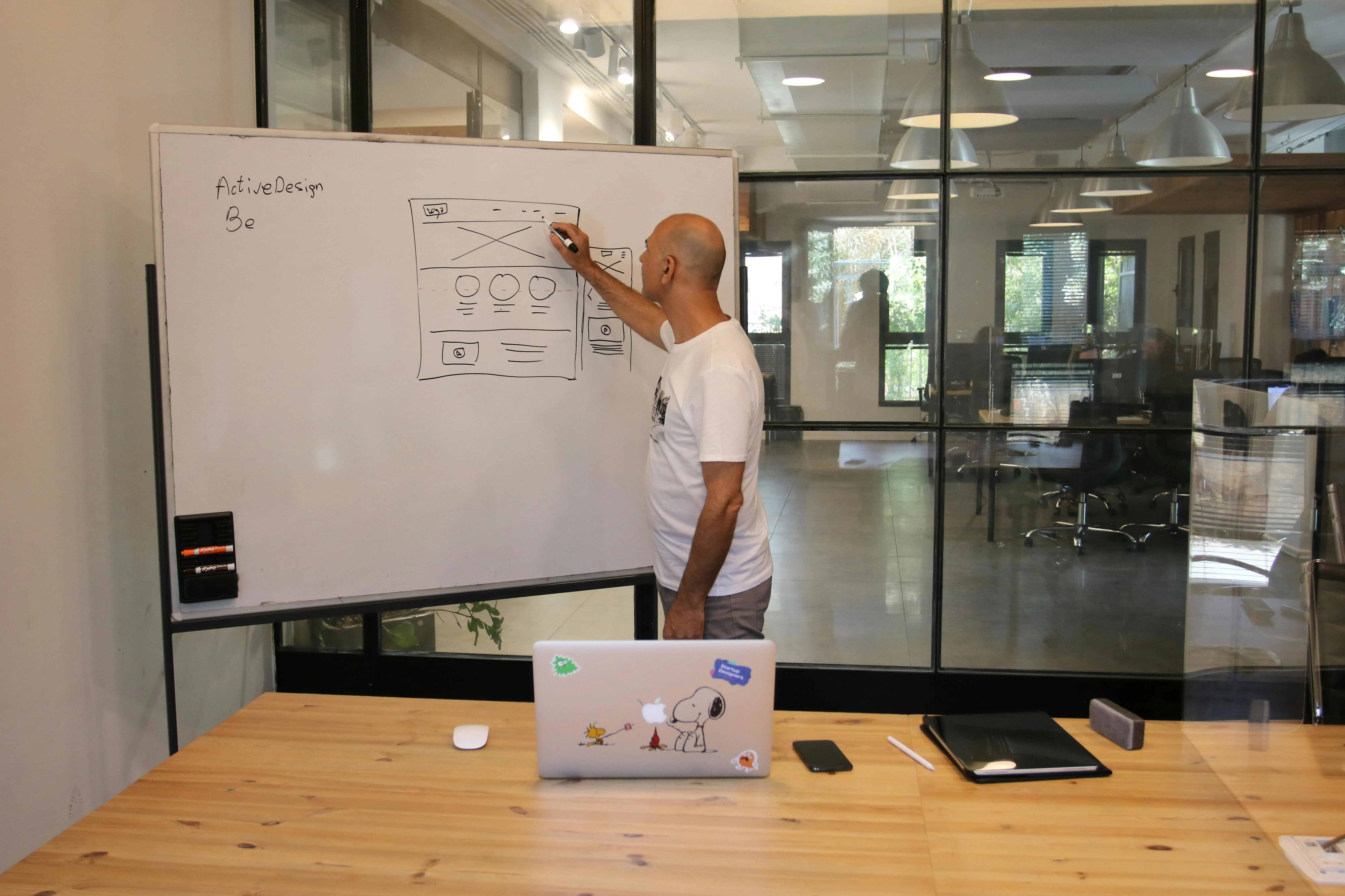 A person sketching a UX Design Process wireframe on a whiteboard labeled "ActiveDesign" in a modern office, with a laptop, notebook, and phone on a wooden table nearby.