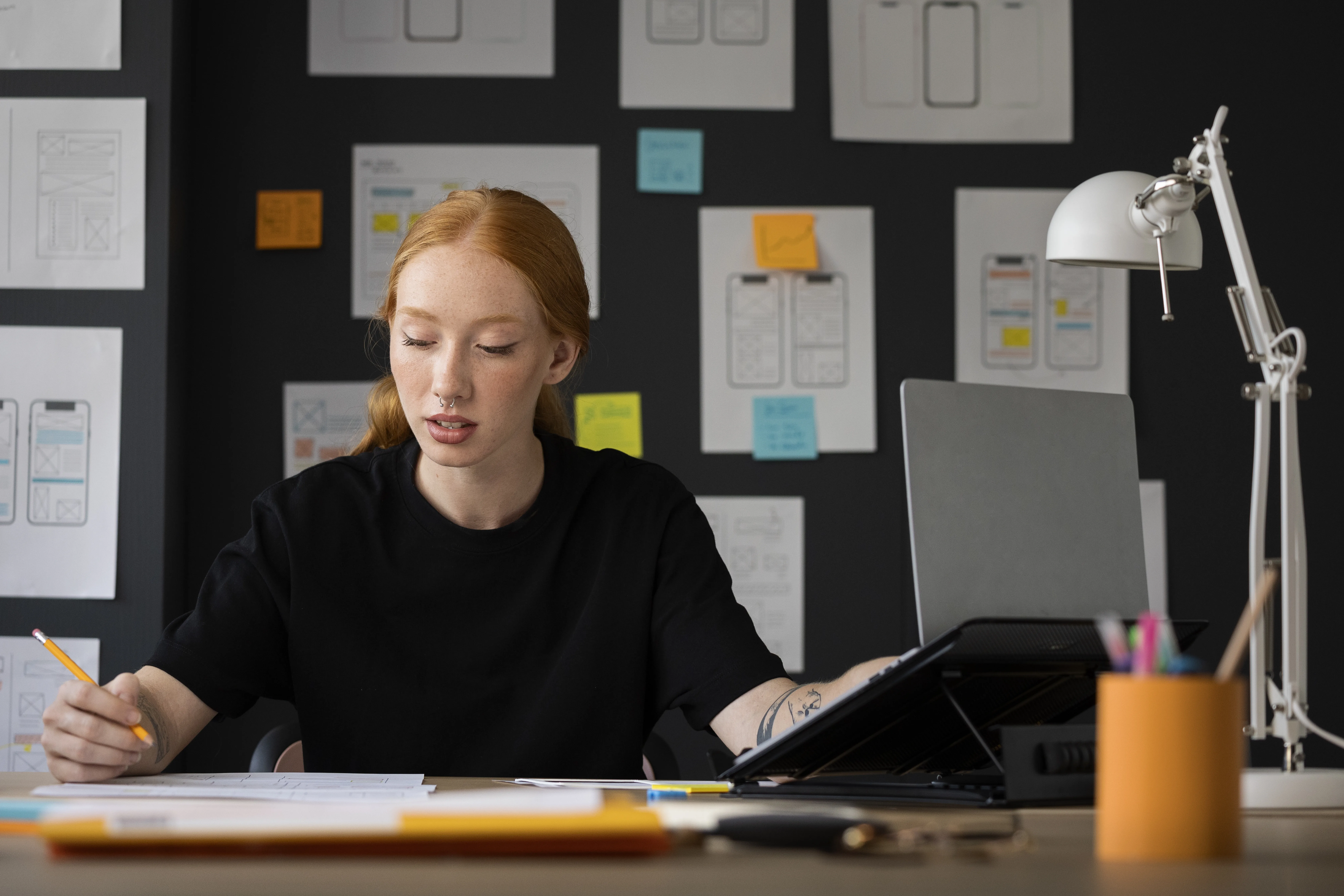 "UX Researcher working at desk with laptop, sketches, and sticky notes on wall, focused on UI/UX design project in creative office setting.