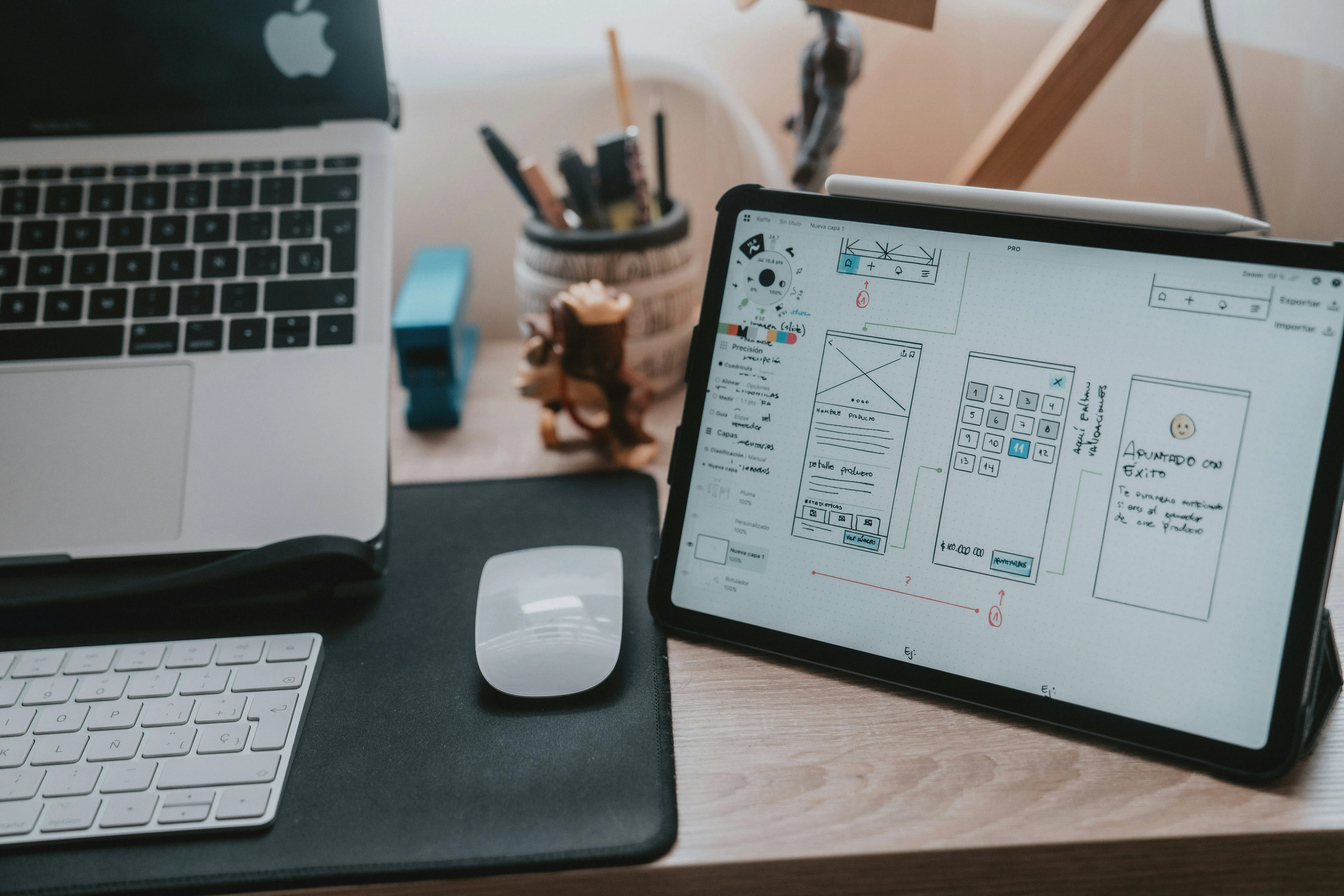 Modern workspace with a MacBook, wireless keyboard, mouse, and tablet displaying a Website Redesign layout, surrounded by office supplies on a wooden desk.