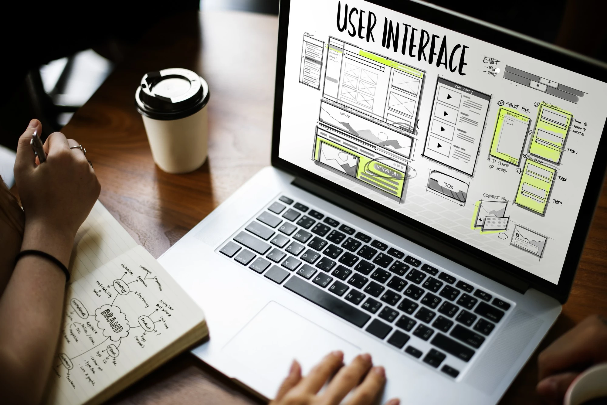 Designer working on Website Redesign user interface on laptop with sketches and coffee cup on wooden table, showcasing creative process.