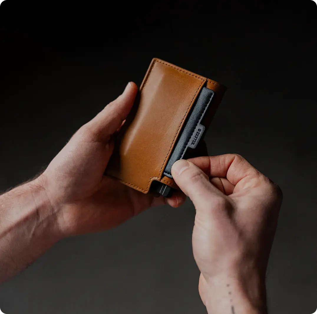 A man's hand pulls a card from a brown leather Ekster wallet with a built-in card-ejection mechanism