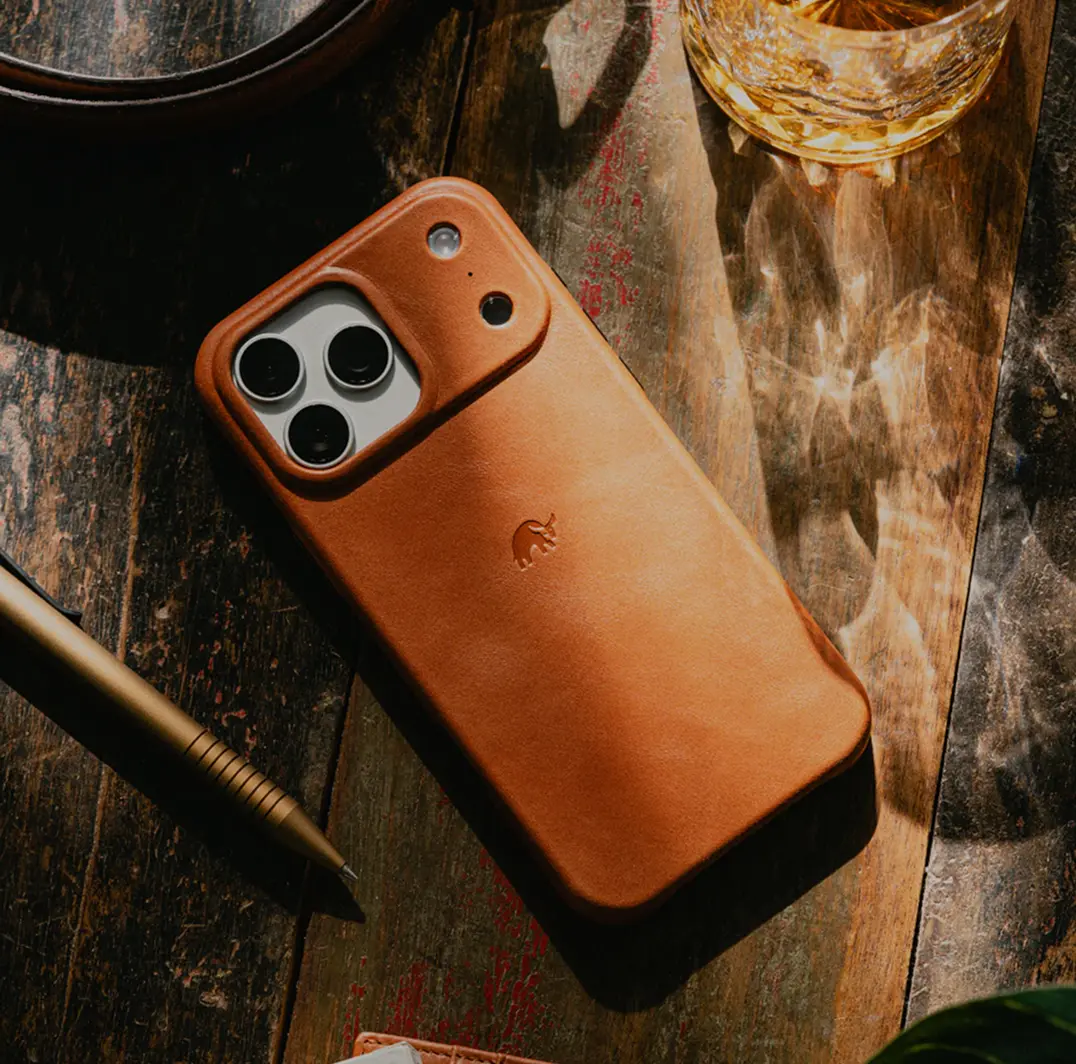 Smartphone in a brown leather Bullstrap case, resting on a wooden surface next to a pen and a glass of drink